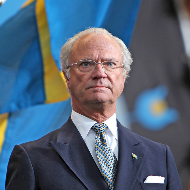 Kungen står framför några svenska flaggor. Han ser allvarlig ut och tittar ut i fjärran. Han har glest vitt hår, glasögon och en kostym med slips.