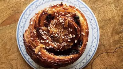 En bulle med pärlsocker och mandel på. Bullen ligger på ett fat med blå kanter. Fatet står på ett yta av grovt trä.