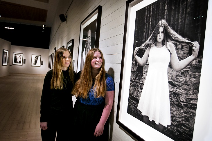 Två personer framför ett foto i ett galleri. I galleriet hänger fotografier. Ett fotografi är på en av personerna som står i galleriet. Personen på fotografiet har en ljus klänning. Personen håller i sitt hår.