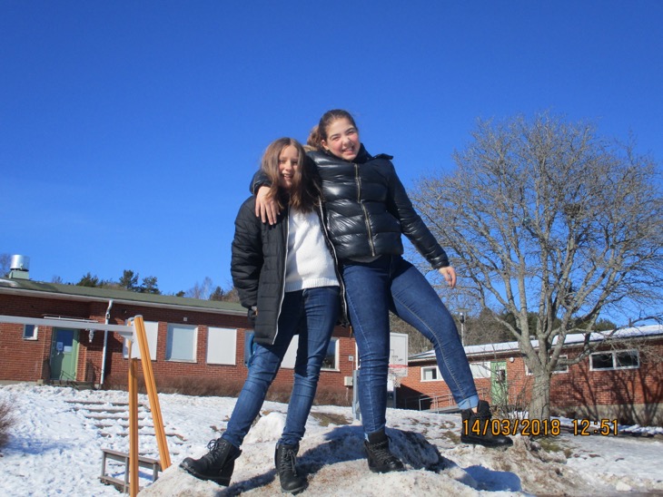 Två barn på en snöhög framför en skola. Himlen är blå. Barnen är glada.