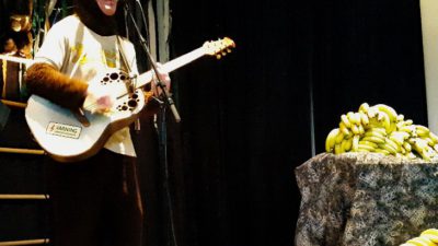 Klasse har på sig en apdräkt och står framför en mick med gitarr. På ett bord intill ligger mängder med bananer.