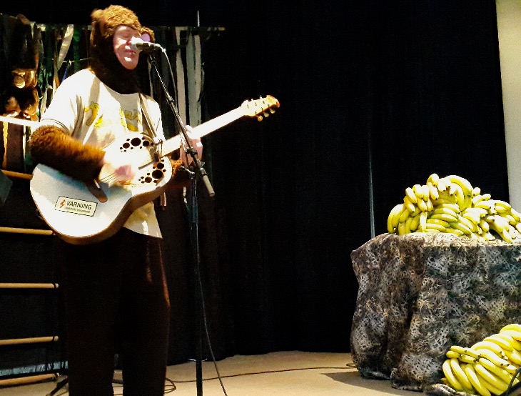 Klasse har på sig en apdräkt och står framför en mick med gitarr. På ett bord intill ligger mängder med bananer.
