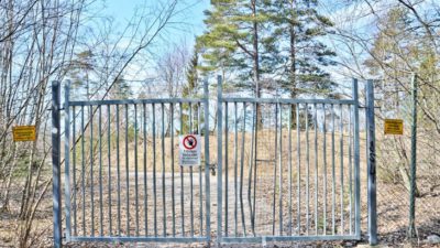 En grind och ett staket med skyltar som säger att det är förbjudet att gå in. Innanför grinden står träd vid sidan om en grusväg.