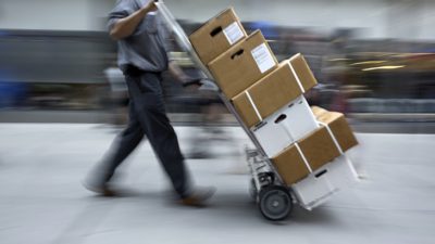 En person går med en vagn framför sig. Vagnen är lastad med lådor som är staplade på varandra.