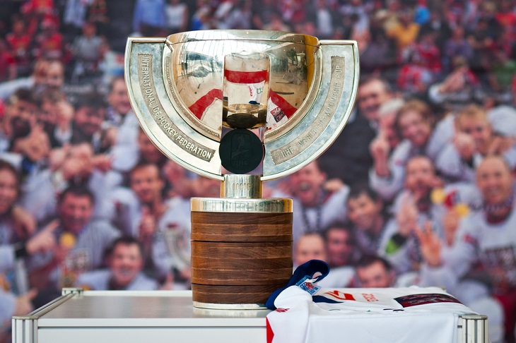 En stor guldig pokal står på ett bort. Pokalen har en fot av trä, en stor skål av guld. Från foten och upp runt var sin sida av skålen går två remsor av metal ut. På remsorna står det text.