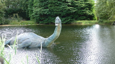 En sjö med träd vid sjökanten. I sjön ligger en stor staty. Statyn har avlång men stor kropp, lång halv, litet huvud och fenor. Den öppnar munnen och gapar mot oss.