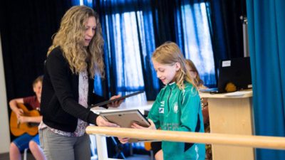 En vuxen håller i två inramade diplom. Barnet tar emot det ena. De är i ett stort rum med stora fönster med fördragna blåa gardiner.