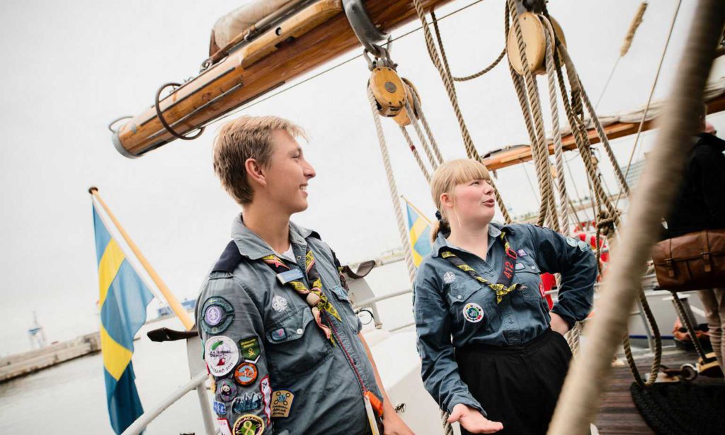 Malcolm och Erik står på däcket på fartyget. Runt dem hänger stora trossar och man ser masten i bakgrunden. De har båda på sig scoutskjortor med massa märken på.